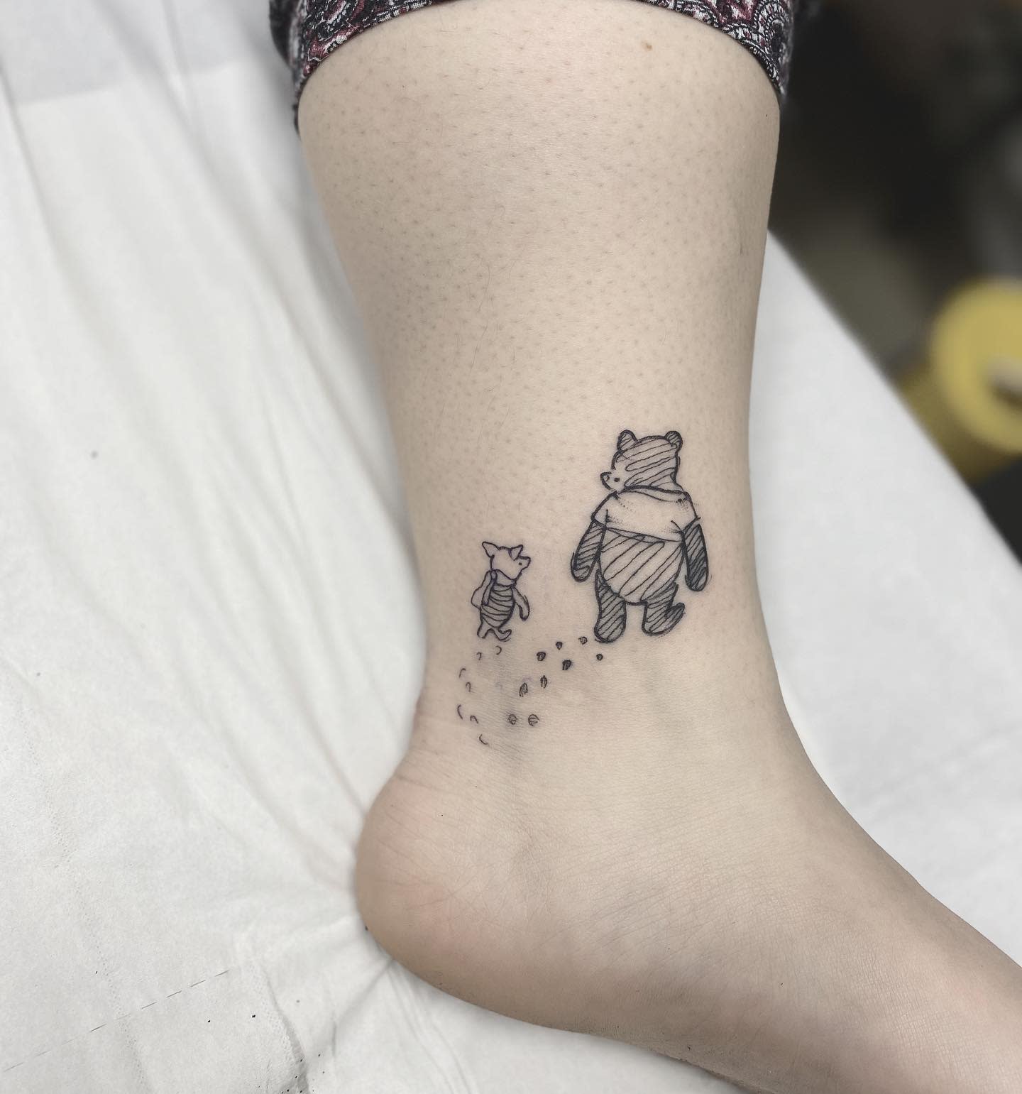 Minimalist tattoo of Winnie the Pooh and Piglet walking together, leaving small footprints, on a person's ankle